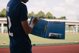 Close up on a soccer player holding a clipboard holding a diagram of the field, the real-world version of which is visible in the background.