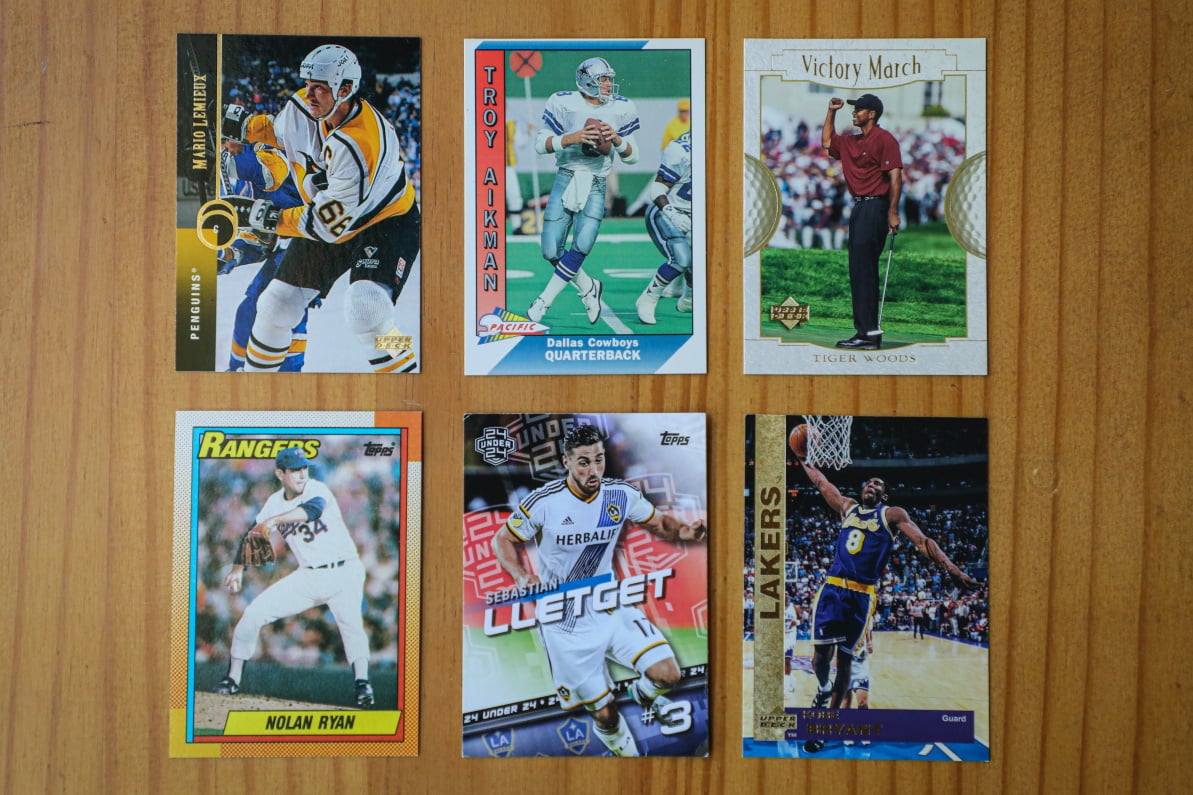 Six sports trading cards featuring Mario Lemieux, Troy Aikman, Tiger Woods, Nolan Ryan, Sebastian Lletget, and Kobe Bryant on a wooden table.