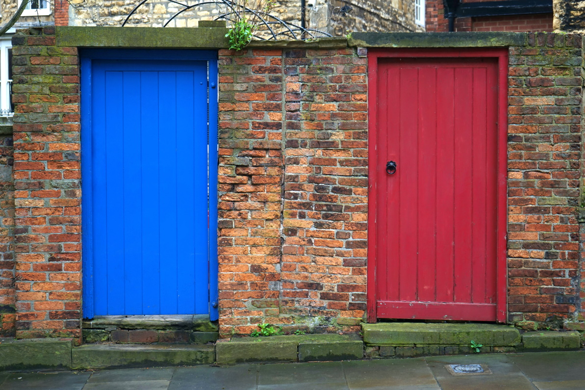 Two wooden doors set into a brick wall. The one on the left is painted bright blue, and the one on the right is red.