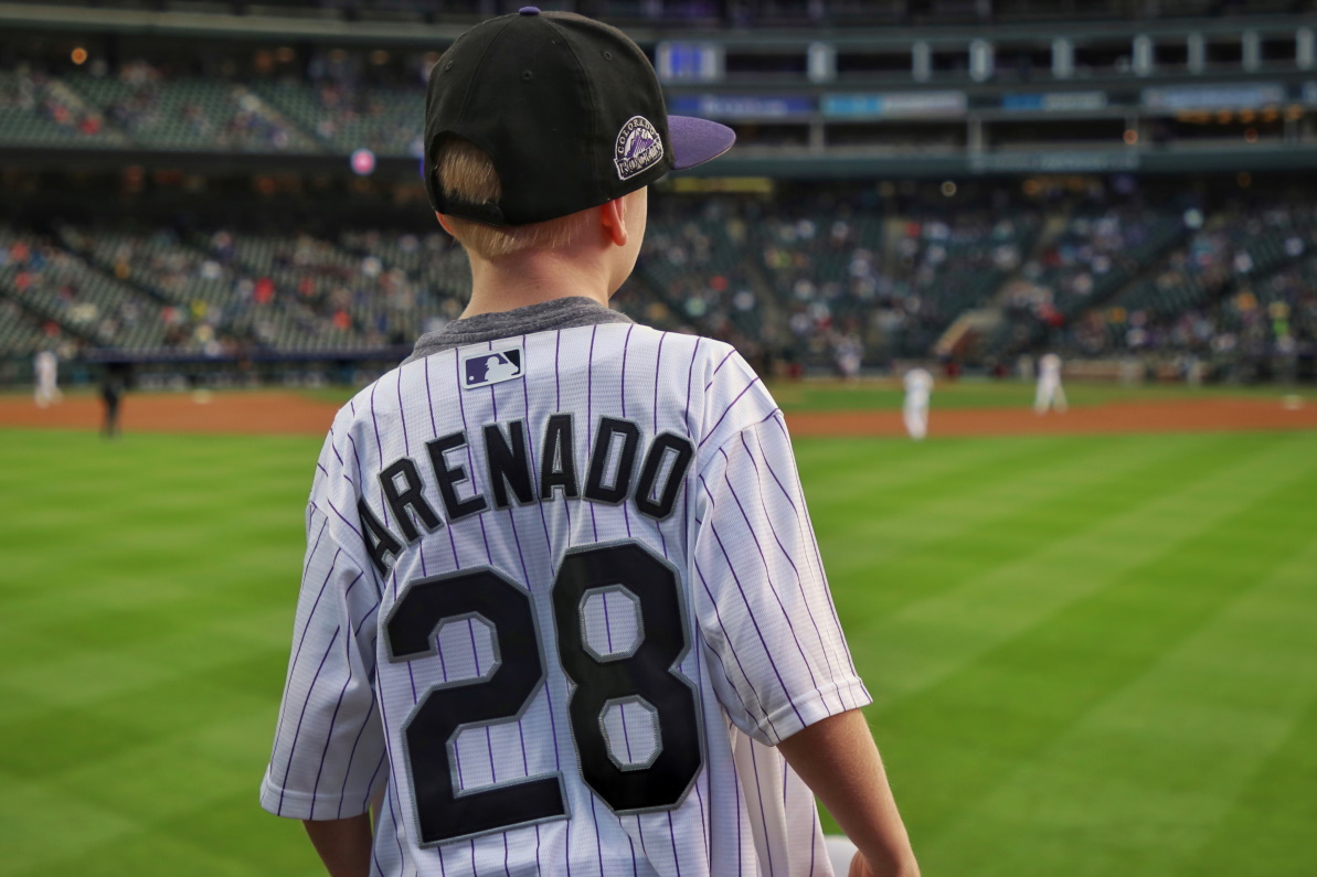 A young boy in a Nolan Arenado jersey and Colorado Rockies hat stands in front of a baseball diamond with his back to the camera.