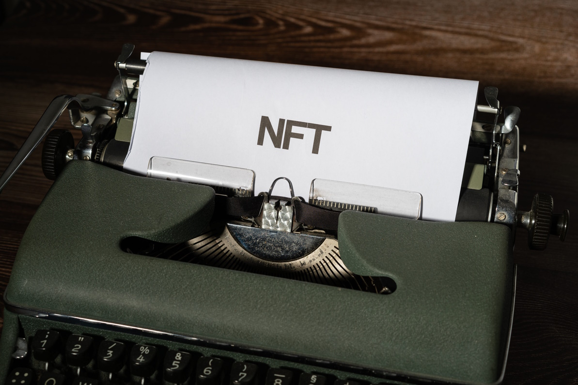 The word 'NFT' is printed on a piece of paper in a bold font. It sits in a green typewriter